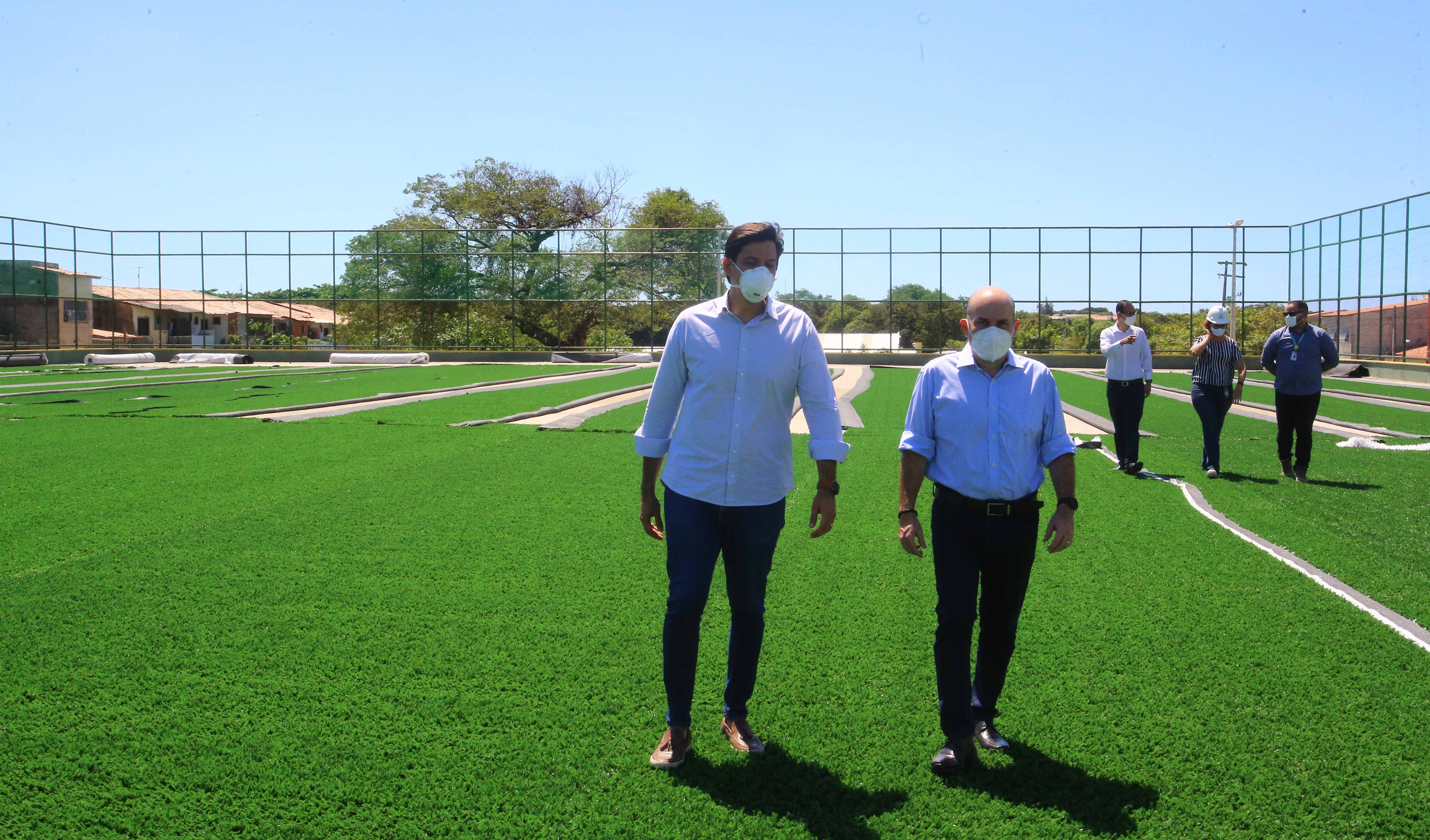 prefeito e secretário caminhando por campo de areninha e três pessoas ao fundo caminhando lado a lado